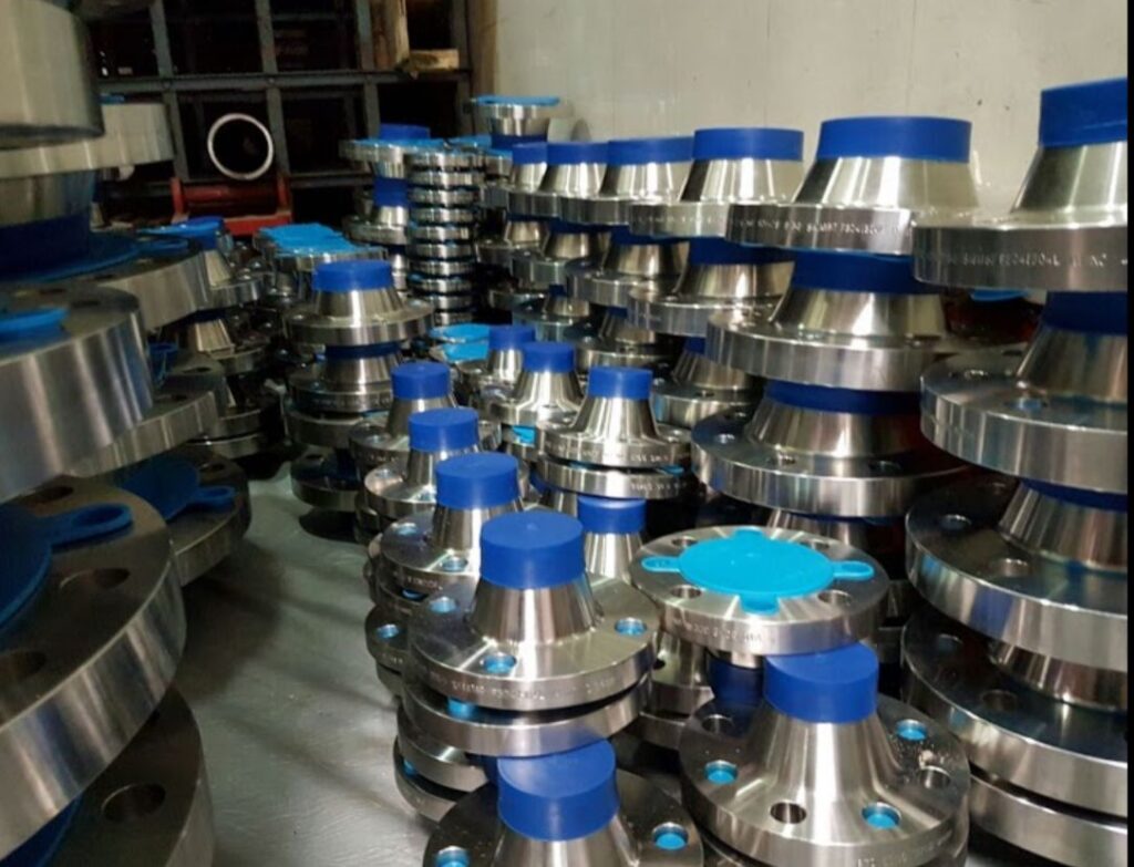 Stainless steel industrial pipe flanges with blue protective caps stacked in a manufacturing factory warehouse.